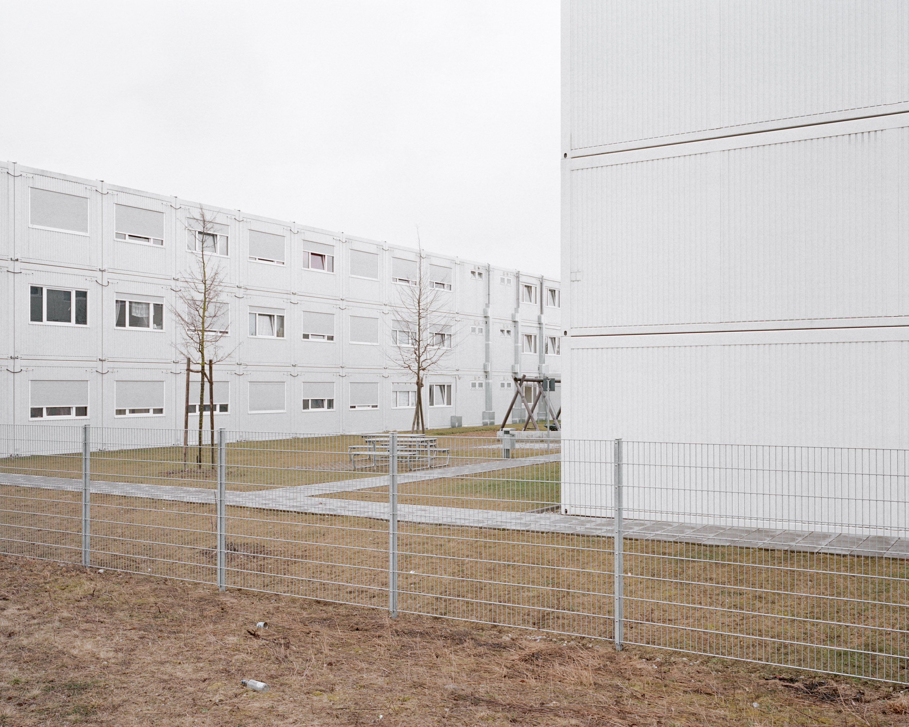 The picture shows the outside of an accommodation for refugees. You see a number of rather greyish buildings and a fence. Its a rather new building, with temporary character.